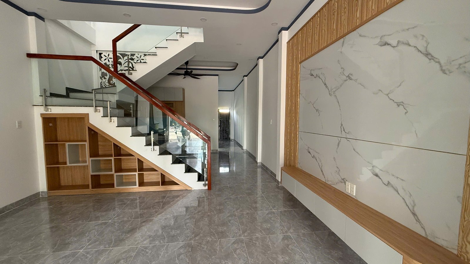 Phòng khách tầng trệt có vách ốp giả đá, cầu thang kính và hộc tủ gỗ dưới gầm cầu thang