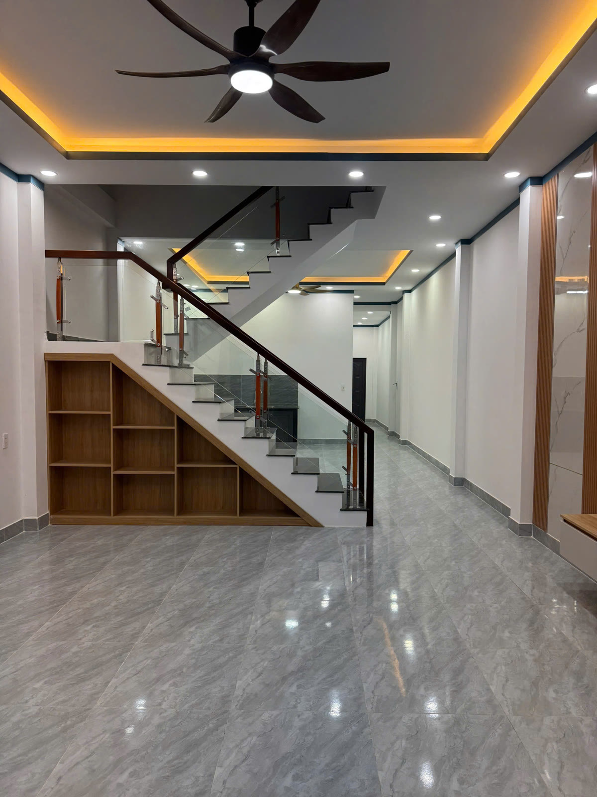 Phòng khách nhà lầu trệt 5x20 Mỹ Phước với trần đèn hắt, quạt trần, cầu thang lan can kính và kệ dưới cầu thang